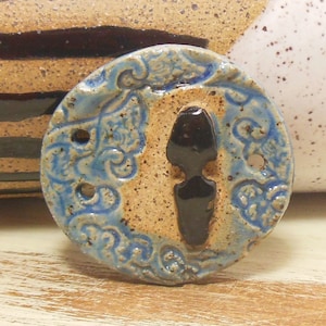 May include: A round ceramic button with a blue and brown glaze. The button features a floral design and a black silhouette in the centre.