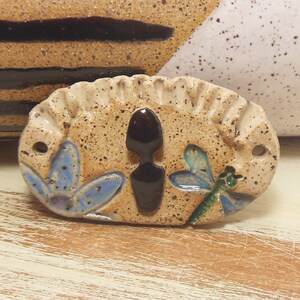 May include: A ceramic oval ornament with a brown silhouette, blue flowers, and a green dragonfly. The ornament has a speckled beige background.