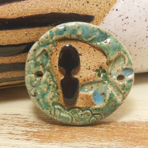 Könnte beinhalten: Eine runde Keramikfliese mit grüner, blauer und brauner Glasur. Die Fliese zeigt eine schwarze Silhouette eines Löffels in der Mitte, umgeben von einem floralen Design.