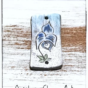 May include: A white ceramic tile with a blue and green floral design. The tile has a hole at the top and is on a white wooden background. The text "Quinlan Glass Art" is at the bottom of the image.