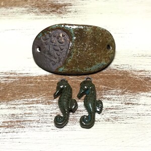May include: Two green ceramic seahorse charms with a light blue glaze. The charms are hanging from a small metal loop. The charms are sitting on a light brown wooden surface.