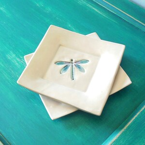 Peut inclure: Deux plats carrés en céramique blanche avec un motif de libellule. Les plats sont empilés l'un sur l'autre et reposent sur une surface peinte en turquoise.