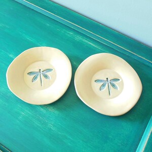 Op de afbeelding: Twee kleine, crèmekleurige keramische schoteltjes met een libellenontwerp. De schoteltjes staan op een turquoise oppervlak.
