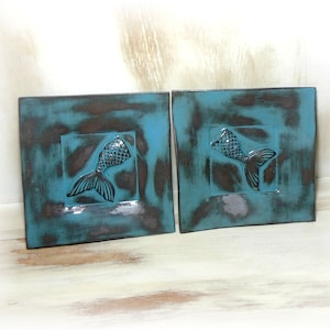 Peut inclure: Deux plaques murales carrées en céramique bleu sarcelle avec un motif en relief de queue de sirène au centre. Les plaques ont un aspect vieilli et vieilli avec des accents bruns et gris.
