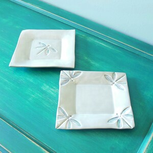 Peut inclure: Deux assiettes carrées en céramique blanche avec des motifs de libellules. Les assiettes sont sur une surface vert turquoise.