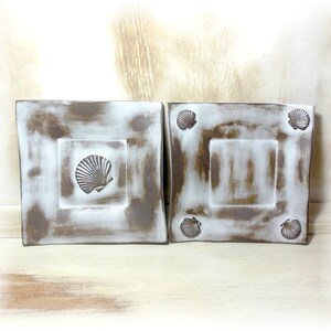 Peut inclure: Deux assiettes carrées en céramique avec un glaçage blanc et des accents bruns. Chaque assiette présente un motif de coquillage en relief au centre et quatre plus petits coquillages autour des bords.