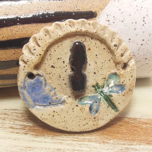 May include: A round, ceramic ornament featuring a brown keyhole, a blue dragonfly, and a blue bird design. The ornament has a speckled, beige background.