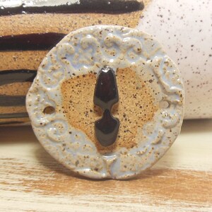 May include: A round ceramic tile with a blue and brown glaze. The tile has a decorative design around the edge and a black, glossy, teardrop-shaped accent in the centre.