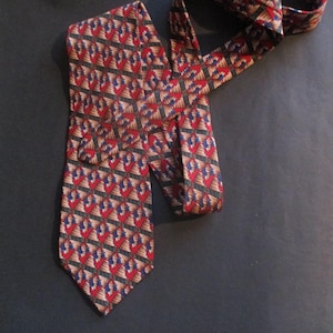May include: A patterned necktie with a repeating design of red, blue, and tan shapes on a dark background. The tie is made of a smooth fabric and is displayed on a dark surface.