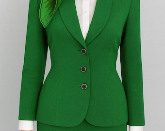 May include: A vibrant green suit with a matching skirt and blazer. The blazer has three black buttons and a classic lapel. A white collared shirt is worn underneath. A green scarf with a leaf pattern is around the neck. The text "The Gourmet Thrifter" is at the bottom.