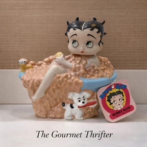 Puede incluir: Una figura de baño de burbujas de Betty Boop. La figura representa a Betty Boop en una bañera con un perro pequeño sosteniendo un cepillo rojo. La etiqueta dice "Betty Boop Bubble Bath". La figura está sobre una superficie blanca.