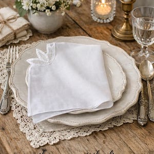 Puede incluir: Una servilleta de lino blanco con un detalle de encaje en la esquina descansa sobre una mesa con platos superpuestos. La mesa está puesta con cubiertos, una vela y un pequeño arreglo floral. La escena evoca una experiencia gastronómica rústica y elegante.