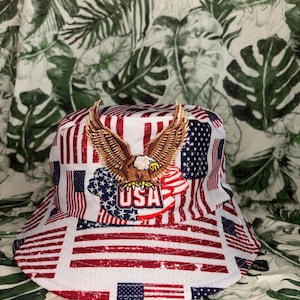 Op de afbeelding: Witte vissershoed met een patriottisch ontwerp, met Amerikaanse vlaggen en het woord "USA". De hoed is rood, wit en blauw, met een geborduurd adelembleem aan de voorkant.