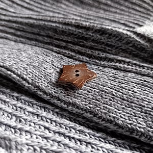 May include: Close-up of a gray knitted fabric with a star-shaped wooden button. The button has two holes and a natural brown color. The fabric has a ribbed texture.