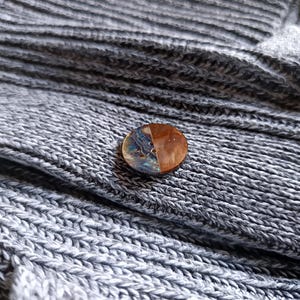 Könnte beinhalten: Ein runder Knopf mit geteiltem Design. Eine Hälfte zeigt eine schillernde, polierte Oberfläche, die andere ist warmbraun. Der Knopf liegt auf einem strukturierten, grauen Strickstoff, was auf ein Kleidungsaccessoire hindeutet.