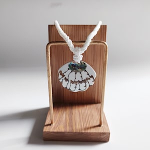 May include: White shell pendant necklace with a white cord. The pendant is a white seashell with a green and blue iridescent centre. The necklace is displayed on a wooden stand.