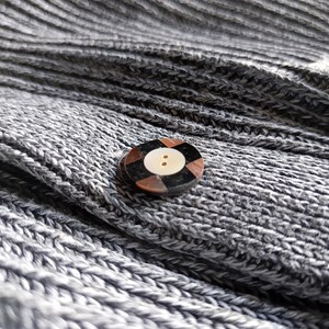 May include: Close-up of a gray knitted fabric with a decorative button. The button is round with a light-colored center and a dark brown and black patterned border. The fabric has a ribbed texture.