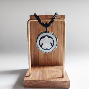 May include: A black beaded necklace with a round, white, mother-of-pearl pendant featuring a carved turtle design. The pendant is surrounded by a black border and is hanging from a wooden display stand.