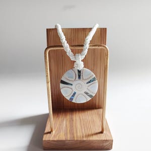May include: A white cord necklace with a round, white and iridescent shell pendant. The pendant has a spiral design and is displayed on a wooden stand.