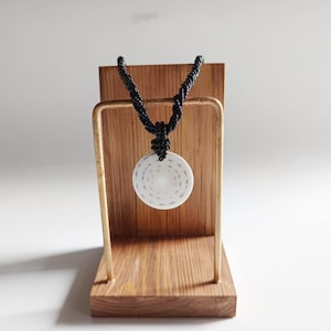 May include: A white ceramic pendant with a spiral design hangs from a black cord necklace. The necklace is displayed on a wooden stand.