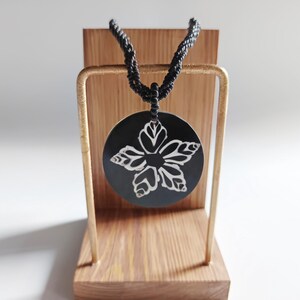 May include: A black necklace with a round pendant featuring a white floral design. The pendant is hanging from a wooden stand.