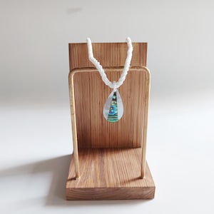 May include: A white beaded necklace with a teardrop-shaped pendant made of iridescent abalone shell. The necklace is displayed on a wooden stand.