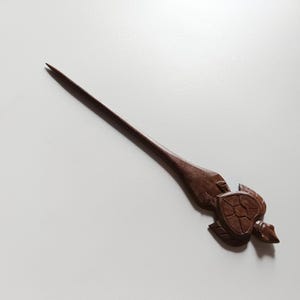 May include: A wooden hair stick with a carved turtle design. The stick is brown and the turtle is detailed with a shell and legs.