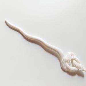 May include: A white hair stick with a wavy stem and a decorative knot design at the end. The hair accessory is made of a smooth, solid material and is set against a white background.
