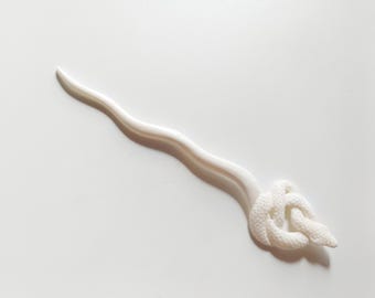 Puede incluir: Una horquilla blanca con un tallo ondulado y un diseño de nudo decorativo en el extremo. El accesorio para el cabello está hecho de un material liso y sólido y se coloca sobre un fondo blanco.