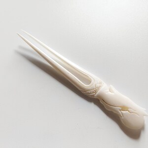 May include: A white carved bone hair fork with a decorative design. The fork has two prongs and a rounded handle.