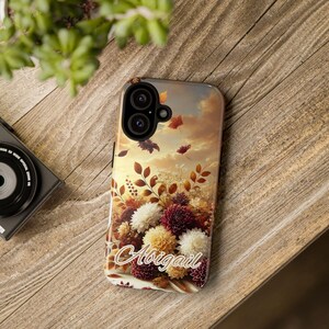 May include: A phone case with a fall-themed design featuring a bouquet of white and red flowers, brown leaves, and a sunset sky. The text "Abigail" is printed on the case.