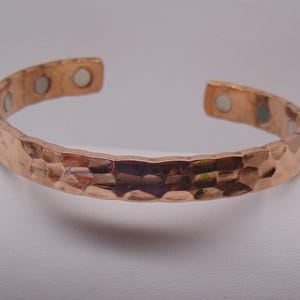 May include: A copper-colored cuff bracelet with a hammered, textured exterior surface. The interior of the open-ended band features several small, circular, silver-toned magnetic inserts embedded at regular intervals along the metal band.