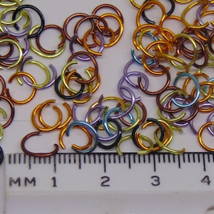 May include: A collection of small, multicolored metal jump rings in shades of gold, copper, blue, purple, and black, scattered above a white plastic ruler. The ruler shows markings in millimeters, with the rings measuring approximately 8 millimeters.