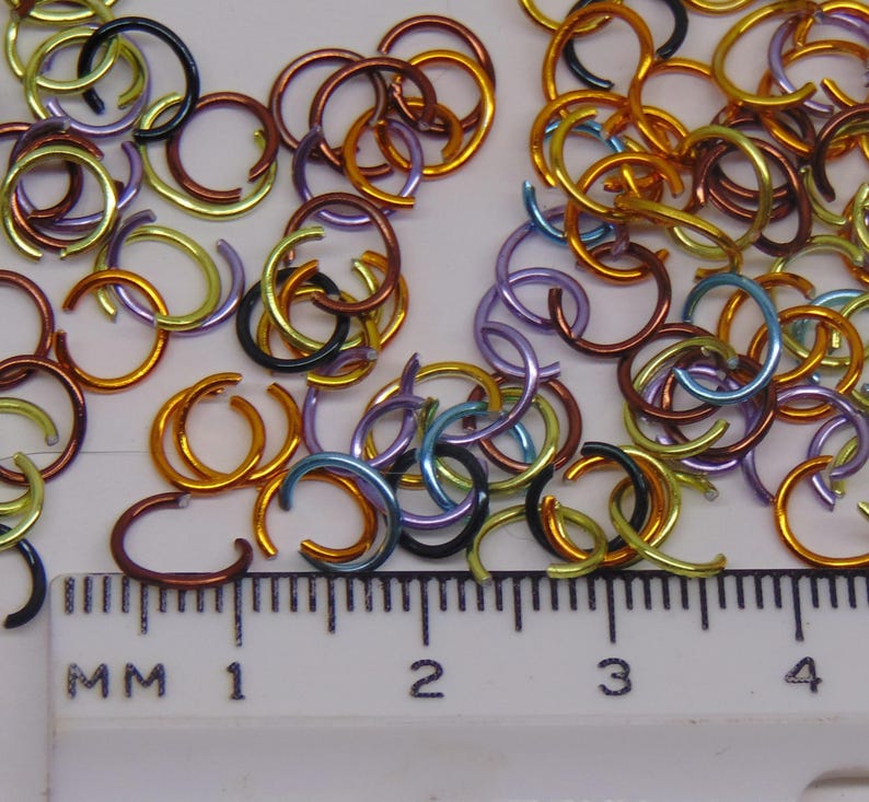 May include: A collection of small, multicolored metal jump rings in shades of gold, copper, blue, purple, and black, scattered above a white plastic ruler. The ruler shows markings in millimeters, with the rings measuring approximately 8 millimeters.