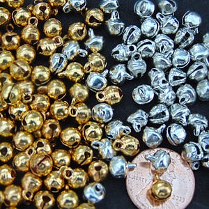 May include: A collection of small, round jingle bells in metallic gold and silver finishes, each featuring a top loop for attachment. A single copper-colored United States penny is positioned among the bells to provide a scale reference for size.
