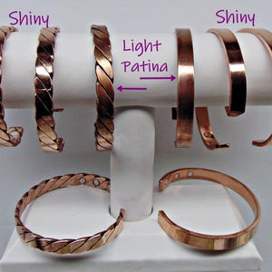 May include: A collection of copper bracelets in various styles. Some are braided, while others are smooth and shiny. The bracelets are displayed on a white stand, showcasing their different designs and finishes. The text "Shiny" and "Light Patina" are visible.