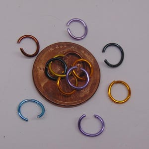 May include: A collection of small, circular metal jump rings in various colors including blue, purple, copper, black, and gold. Several rings are scattered around a United States copper penny, while others are layered directly on top of the coin.