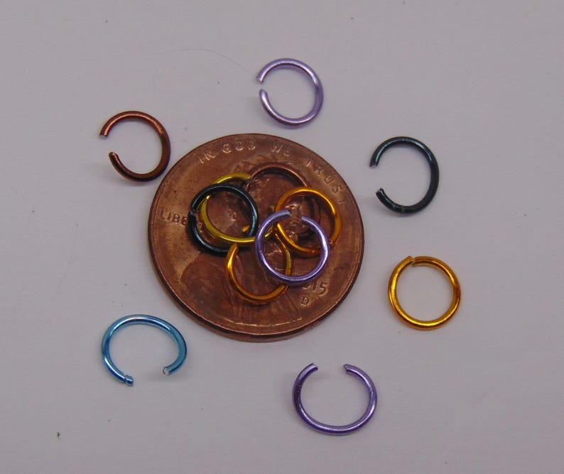 May include: A collection of small, circular metal jump rings in various colors including blue, purple, copper, black, and gold. Several rings are scattered around a United States copper penny, while others are layered directly on top of the coin.