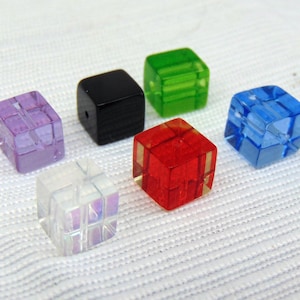 May include: A collection of colorful, translucent cube-shaped beads. The beads are in various colors, including purple, black, green, red, blue, and iridescent white. The cubes are arranged on a white, textured surface. These beads are suitable for jewelry making.