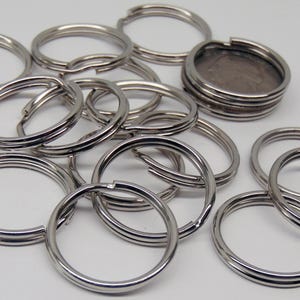 May include: A collection of silver-colored split key rings. The key rings are round with a split opening for attaching keys. Some rings are open, while others are closed. The rings are made of metal and are arranged on a white surface.