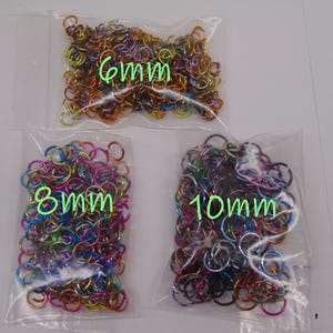May include: Three clear plastic bags filled with multicolored metal jump rings. The bags are labeled with green text indicating sizes of 6mm, 8mm, and 10mm. The rings feature a vibrant, iridescent finish in shades of blue, purple, pink, and gold.