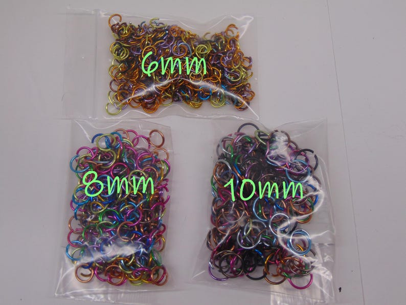 May include: Three clear plastic bags filled with multicolored metal jump rings. The bags are labeled with green text indicating sizes of 6mm, 8mm, and 10mm. The rings feature a vibrant, iridescent finish in shades of blue, purple, pink, and gold.