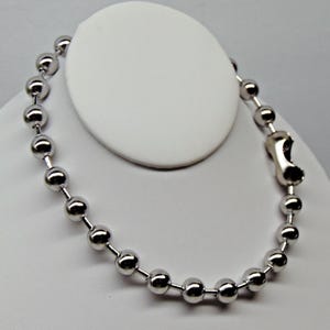 May include: A silver-colored ball chain necklace with a clasp. The necklace is composed of numerous small, round, metallic beads strung together. The clasp is a simple, functional design. The necklace is displayed on a white surface.