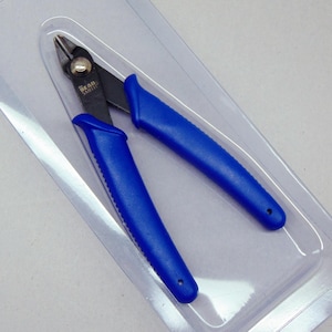 May include: A pair of blue-handled side cutters with black metal jaws, featuring the text "THE BEAD SMITH" printed on the metal. The tool is encased in a clear plastic blister pack, showcasing the ergonomic blue grips and the sharp cutting edges.