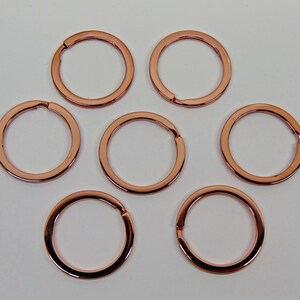 May include: Seven copper-colored split key rings are arranged on a white surface. Each ring is circular with a small opening for attaching keys or other items. The rings have a shiny, metallic finish.