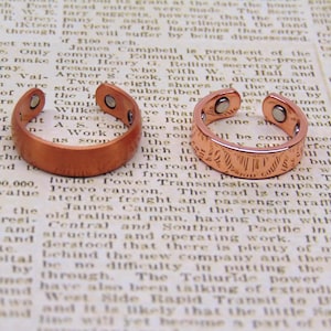 May include: Two open-ended copper-toned rings with a smooth, polished finish. Each ring features two small, circular silver-colored magnets embedded on the interior surface at both ends of the band, designed for a flexible, adjustable fit.