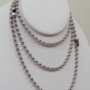 May include: A layered necklace with three strands of small, silver-colored ball chain. The necklace is displayed on a white bust form. Each strand is connected with a silver clasp. The necklace is a simple, yet elegant accessory.