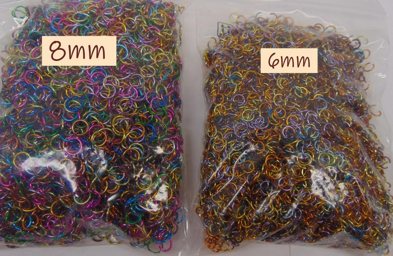 May include: Two clear plastic bags filled with small metal jump rings. The left bag contains multicolored rings labeled 8mm, while the right bag contains gold and bronze-toned rings labeled 6mm. These circular metal components are used for jewelry making.