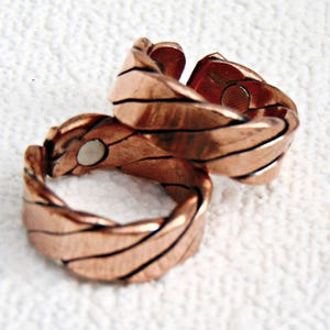 May include: Two copper-colored rings with a textured, woven design. Each ring features a series of curved, parallel lines and a small, circular magnet. The rings are displayed on a white, textured surface.