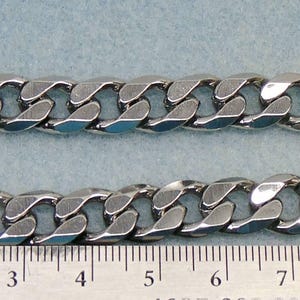 May include: Two silver-colored curb chains are displayed side-by-side. The chains have a series of interlocking, rounded links. A ruler with millimeter markings is at the bottom, providing a size reference. The chains are on a light blue background.